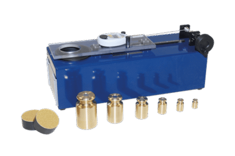 The Surface Shear Test Apparatus features a blue mechanical device with a metal dial, sliding mechanism, six brass weights of assorted sizes, and two round black and yellow pads, displayed on a white background.
