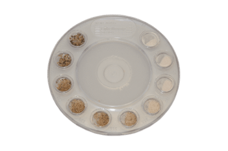 The Sand Ruler is a round plastic tray with ten small compartments, each holding a unique sand or soil sample, arranged in a circle on a white background.